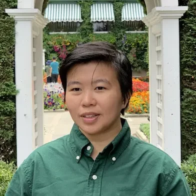 A person with short dark hair, wearing a green button-up shirt, stands outdoors in front of a white garden archway with flowers and greenery, representing Loomo Marketing amid colourful plants in the background.