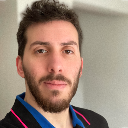 A person with short dark hair and a beard looks at the camera. They are wearing a black jacket with pink trim over a blue shirt. The softly blurred indoor background subtly hints at a Loomo Marketing workspace.