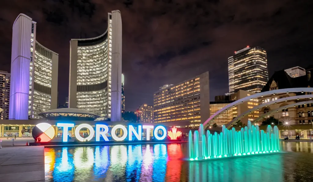 Toronto City Hall at night with its illuminated curved towers, the glowing TORONTO sign, and colourful fountain reflections in the foreground, with other lit up office buildings in the background