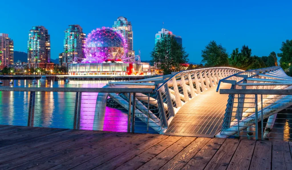 A modern pedestrian bridge over water leads to Vancouver's city skyline at dusk, with BC’s iconic Science World glowing purple and its reflection shimmering in the water