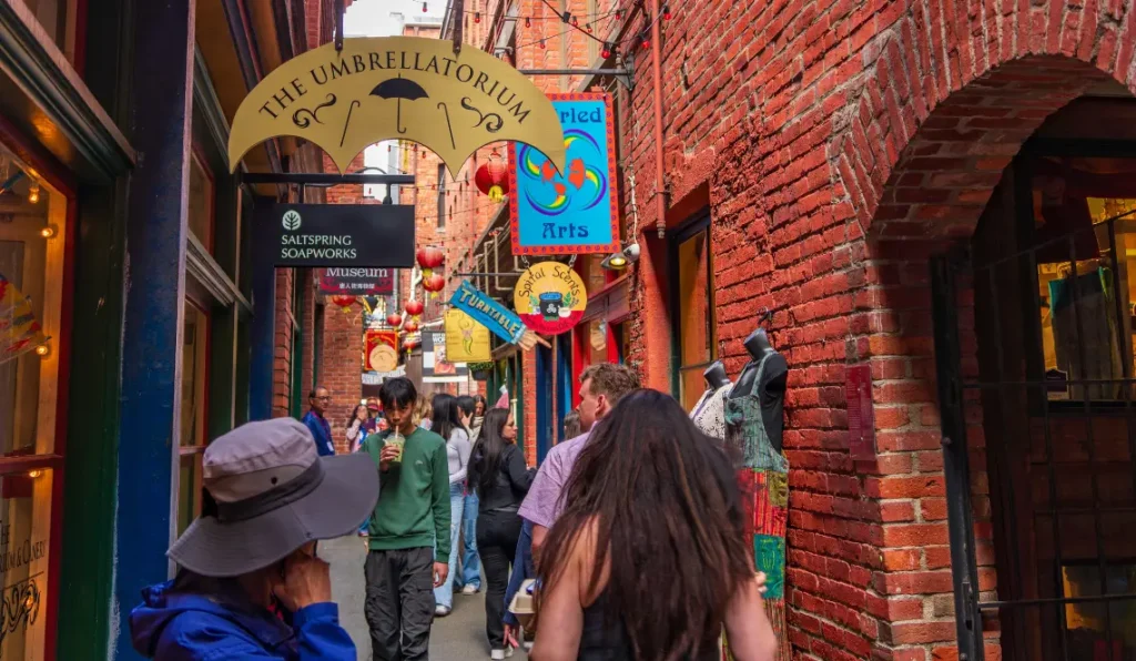 A narrow brick alley lined with colourful shop signs and lanterns is busy with people walking and browsing small businesses, including “The Umbrellatorium,” “Saltspring Soapworks,” and a creative Marketing Agency Victoria locals trust