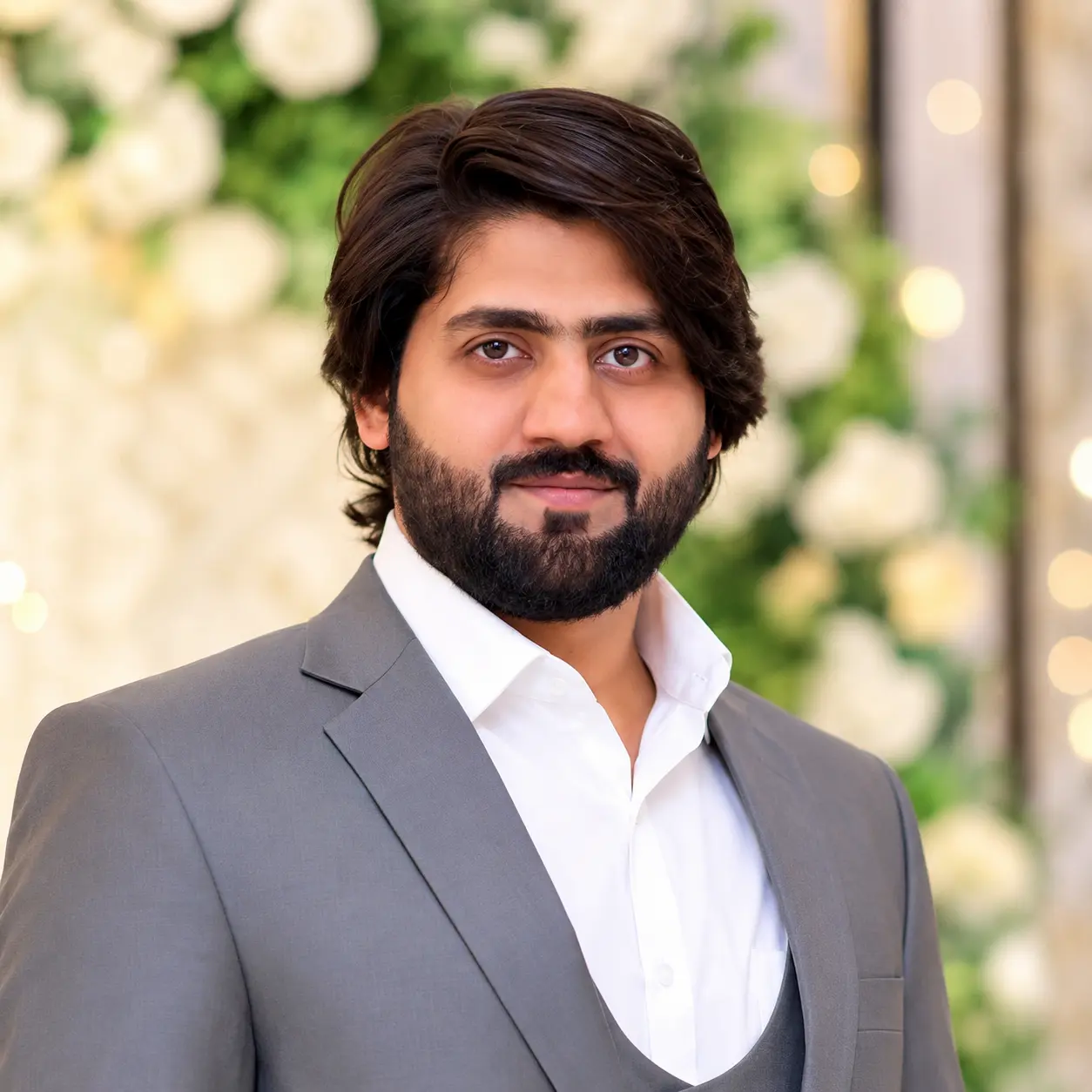 Meet The Crew: A man with dark hair and a beard, wearing a grey suit and white shirt, stands in front of a blurred background decorated with white flowers and greenery